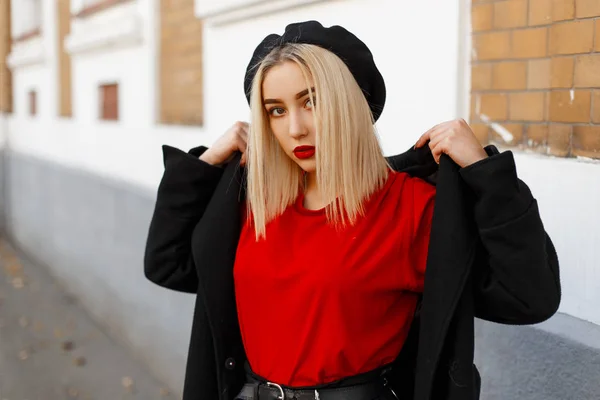 Loira jovem moderna com um penteado com lábios vermelhos sexy em uma boina preta elegante em um casaco longo e elegante em uma camiseta vermelha posando perto de um prédio antigo. Muito elegante menina moda model.Youth estilo — Fotografia de Stock