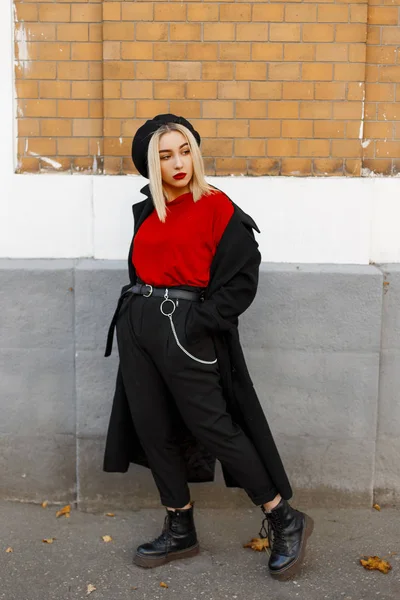 Αρκετά κομψή νεαρή ξανθιά γυναίκα σε ένα μοντέρνο παλτό σε μοντέρνα παντελόνια σε ένα κόκκινο πουκάμισο σε ένα μπερέ σε δερμάτινες μπότες αναπαύεται στέκεται κοντά σε ένα vintage τούβλο τοίχο μια ημέρα του φθινοπώρου. Δροσερό κορίτσι σε εξωτερικούς χώρους. — Φωτογραφία Αρχείου