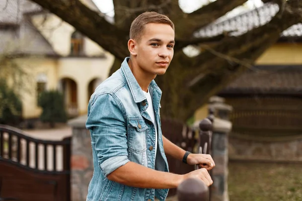 Beyaz bir t-shirt bir moda denim ceket şık çekici adam ayakta ve evin yakınındaki bahçesinde bir Vintage Ferforje çit yakın gülümseyen. Sevimli şık Amerikan adam. Bahar tarzı.