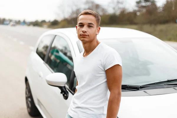Young handsome sporty man with fashionable hairstyle in trendy white t-shirt stands near a white car outdoors. Stylish attractive guy traveling by car.