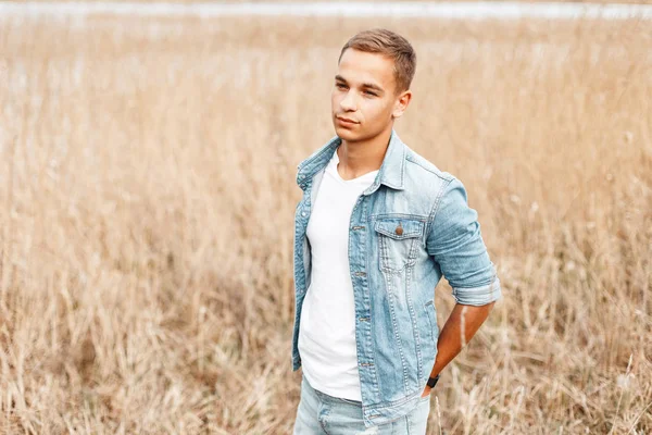 Handsome cute young man in a fashionable denim jacket in a stylish white t-shirt in vintage blue jeans is standing in a field among the dry grass. Attractive modern guy enjoys nature.