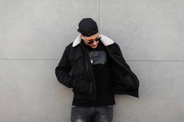 Siyah beysbol şapkalı, güneş gözlüklü, modaya uygun siyah ceketli, gri kotlu beyaz kürklü, modern bir binanın yanında duran havalı bir hippi. Yürüyüş için yakışıklı şehir çocuğu modeli.