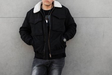 Siyah renkli ceketli, beyaz yakalı, siyah tişörtlü, modaya uygun genç bir adam gri bir duvarın yanında duruyor. Modaya uygun erkek kıyafetleri. Bir erkek cesedine yakın çekim.