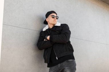 Gri vintage kot siyah şık bir ceket trendy Amerikan beyzbol şapkası güneş gözlüğü Çekici serin genç adam hipster modern gri binanın yakınında dinleniyor. Yürüyüş için yakışıklı adam.