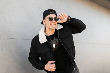 Güneşli bir günde gri bir duvarın yakınında poz güneş gözlüğü beyaz kürk lü moda bir ceket modat shirt şık beyzbol şapkası Pozitif yakışıklı genç adam hipster. Neşeli adam bir yürüyüş zevk.
