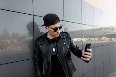 Bir t-shirt trendy deri ceket güneş gözlüğü şık kap Yakışıklı serin genç hipster adam modern gri ayna bina yakınında bir selfie yapar. Şehirde sokakta çekici adam.