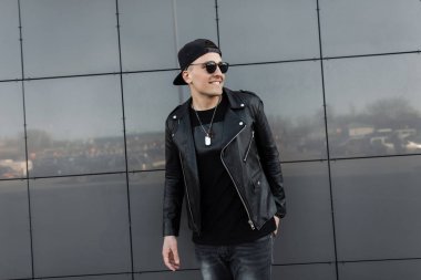 Trendy bir kap güneş gözlüğü bir t-shirt bir vintage deri ceket olumlu mutlu genç hipster adam modern bir gri ayna bina yakınında duruyor ve hafta sonu zevk. Neşeli adam.