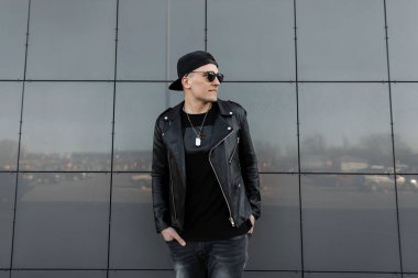 Güneş gözlüğü siyah bir beyzbol şapkası şık siyah giysiler içinde Modern moda genç hipster adam sıcak bir bahar gününde gri binanın yanında duran rahatlatır. İyi adam. Trendy erkek giyim.