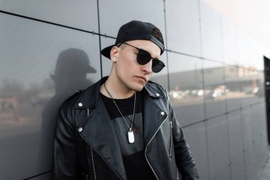 Şık bir t-shirt şık bir deri ceket güneş gözlüğü siyah bir kap çekici genç hipster adam modern parlak alışveriş merkezi yakınında dinleniyor. Şehirde sokakta Cool adam.