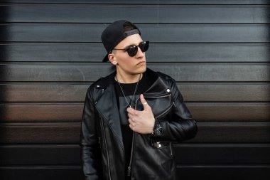 Modaya uygun bir beyzbol şapkası siyah deri ceket moda koyu güneş gözlüğü Güzel genç hipster adam modern bir ahşap siyah evin yakınında poz. Sokaktaki yakışıklı adam. Retro tarzı.