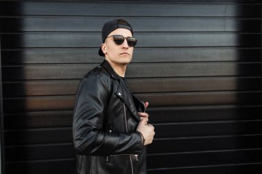 Modaya uygun siyah güneş gözlüğü bir moda siyah beyzbol şapkası bir vintage deri ceket Ciddi şık genç hipster adam modern bir ahşap siyah evin yakınında duruyor. Sokakta Amerikalı adam. 