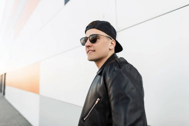 Koyu moda güneş gözlüğü bir deri şık ceket şık siyah kap Mutlu Amerikan hipster genç adam ayakta ve karanlık bir bahar gününde modern duvarın yakınında gülümsüyor. Neşeli adam modeli.
