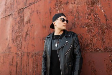 Siyah beysbol şapkalı, siyah güneş gözlüklü, gri kotlu, modaya uygun siyah ceketli, dışarıda paslı metal bir duvarın yanında dinlenen genç ve çekici bir hippi. Yürüyüş için yakışıklı bir adam..