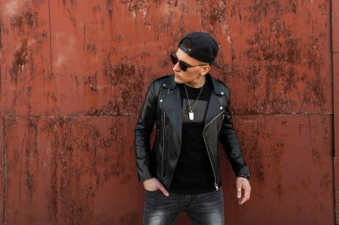 Gri kot bir vintage T-shirt bir kap şık deri siyah ceket güneş gözlüğü ile Modern moda genç hipster adam bir yaz gününde metalik kırmızı binanın yakınında duruyor. Yakışıklı adam.