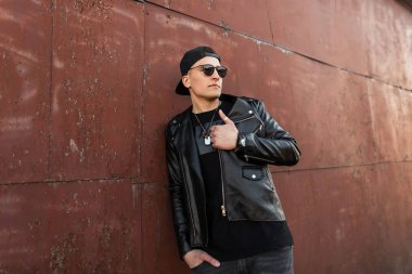 Bir yaz gününde metal bir duvarda poz gri kot moda siyah t-shirt siyah şık bir kap koyu güneş gözlüğü bir deri moda ceket yakışıklı genç hipster adam. Cool adam dinlenme.