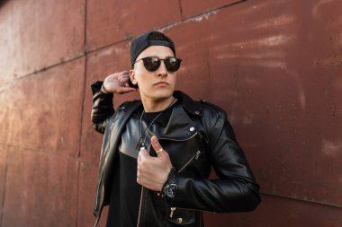 Siyah bir t-shirt koyu moda güneş gözlüğü şık bir deri siyah ceket yakışıklı genç adam hipster bir vintage metal duvar açık yakınında duruyor. Çekici erkek modeli.