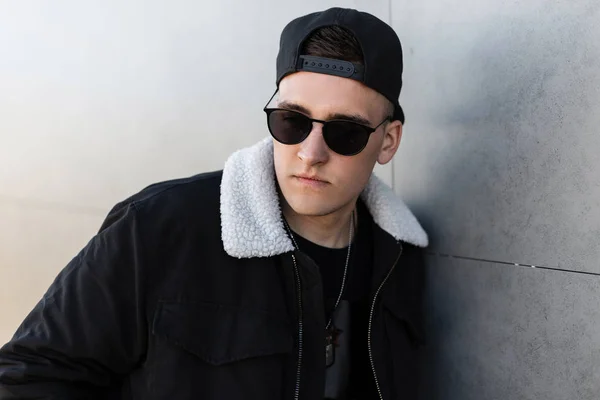 Guapo joven hipster en una elegante chaqueta negra en una gorra negra de moda en gafas de sol de moda oscura en una camiseta negra se encuentra cerca de la moderna pared gris al aire libre. Muchacho atractivo modelo de moda . — Foto de Stock