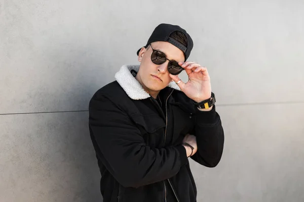 Young attractive hipster man in a trendy baseball cap in stylish sunglasses in a vintage black jacket with a white collar poses for a spring day in the city near a modern building. American guy. — Stock Photo, Image
