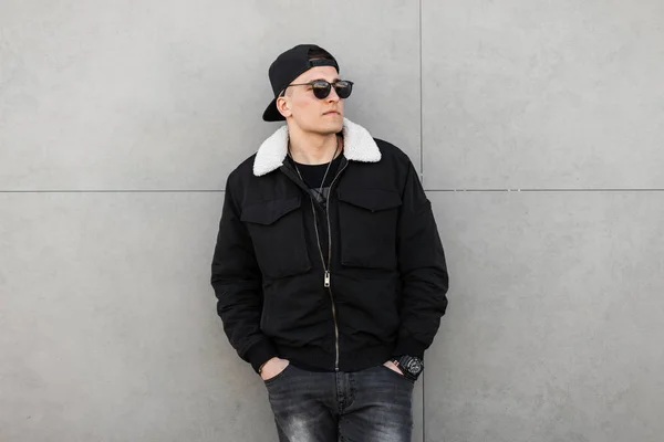 Young attractive hipster man in a black baseball cap in a dark sunglasses in a trendy black jacket with white fur in gray jeans standing near a modern building. Handsome city guy model for a walk.
