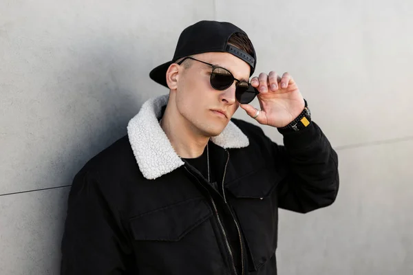 Joven hombre hipster americano de moda en gafas de sol con estilo en una elegante gorra de béisbol negro en una chaqueta de moda negro posa cerca de la pared moderna en un día de primavera. Chico elegante descansando al aire libre . — Foto de Stock