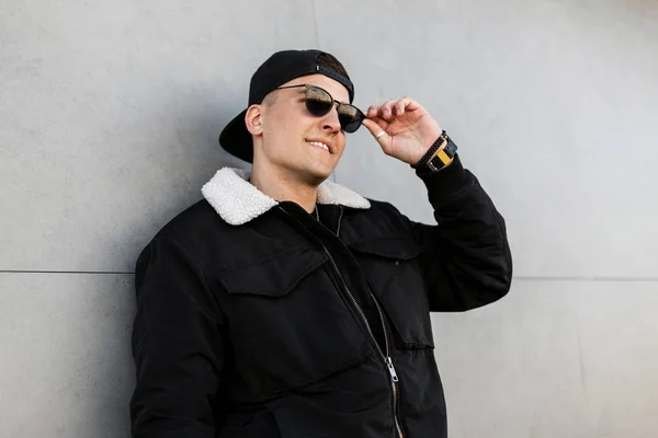 Un joven con estilo positivo hipster en una gorra de béisbol negra con gafas de sol en una chaqueta de moda se relaja al aire libre cerca de un edificio moderno en un día de primavera. Un tipo feliz disfrutando el fin de semana. Buenos días. . — Foto de Stock