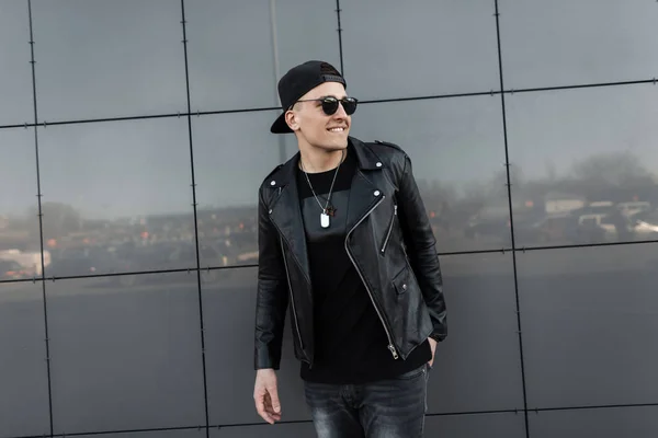 Positivo joven hipster feliz hombre en una chaqueta de cuero vintage en una camiseta en jeans con gafas de sol en una gorra de moda está de pie cerca de un moderno edificio de espejo gris y disfrutar del fin de semana. Chico alegre . — Foto de Stock