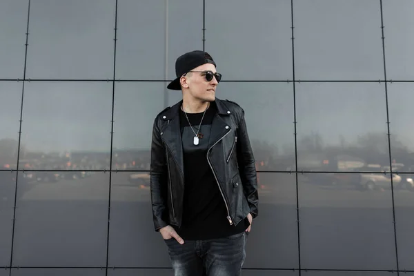 Moderno hombre hipster joven de moda en ropa negra con estilo en una gorra de béisbol negro en gafas de sol se relaja de pie cerca del edificio gris en un cálido día de primavera. Buen tipo. Ropa de hombre de moda . — Foto de Stock