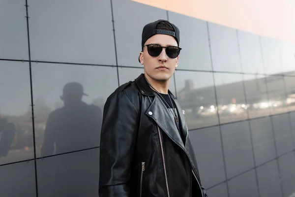 Elegante joven guapo hipster en gorra de béisbol de moda en chaqueta de cuero negro vintage con gafas de sol con estilo en una camiseta se encuentra cerca de una moderna pared de espejo gris al aire libre. Buen tipo descansando. . — Foto de Stock