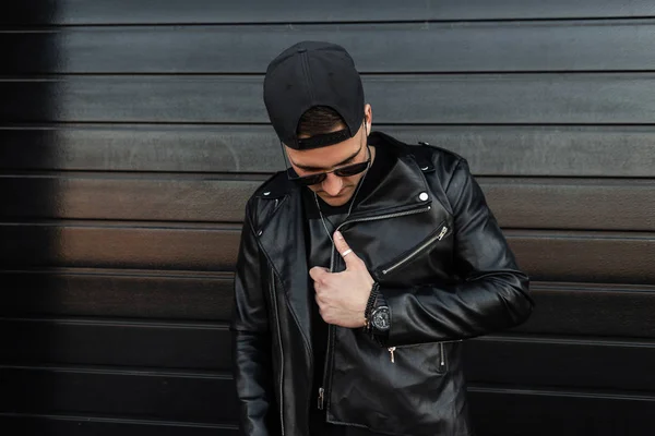 Fashionable young hipster man in sunglasses in a vintage leather jacket in a stylish baseball cap relaxes near a modern wooden black wall. Handsome guy in the street. Nice guy. Youth style clothing.
