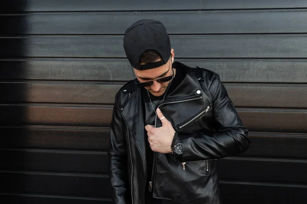 El joven hipster de moda con gafas de sol en una chaqueta de cuero vintage con una elegante gorra de béisbol se relaja cerca de una moderna pared negra de madera. Un tipo guapo en la calle. Buen tipo. Ropa de estilo juvenil . — Foto de Stock