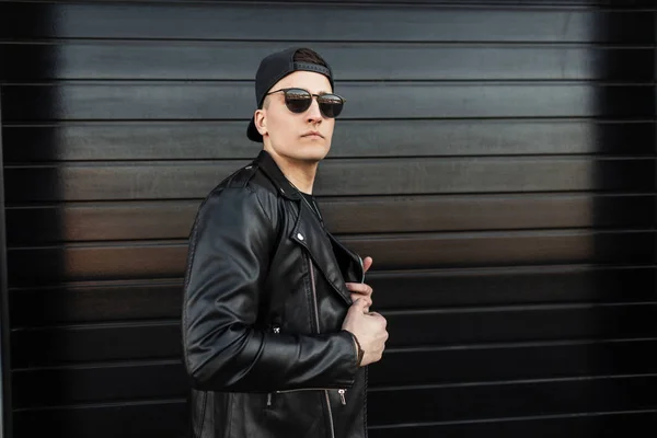 Serious stylish young hipster man in a vintage leather jacket in a fashionable black baseball cap in a trendy dark sunglasses is standing near a modern wooden black house. American guy on the street. — Stock Photo, Image