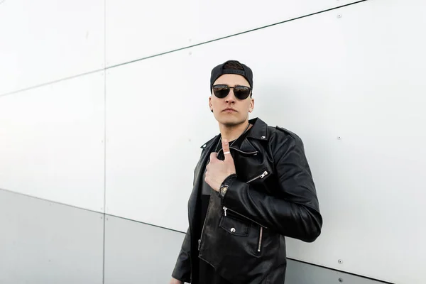 Attractive stylish young hipster man in stylish leather jacket in trendy black baseball cap in trendy dark sunglasses poses near modern white building in the city. American guy on the street. Retro.