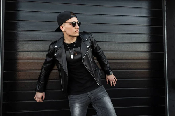 Joven hombre hipster fresco en elegante camiseta en una gorra de béisbol negro de moda con gafas de sol oscuras en una chaqueta de cuero negro de moda en jeans grises posando junto a una pared de madera negra. Guapo chico al aire libre . — Foto de Stock