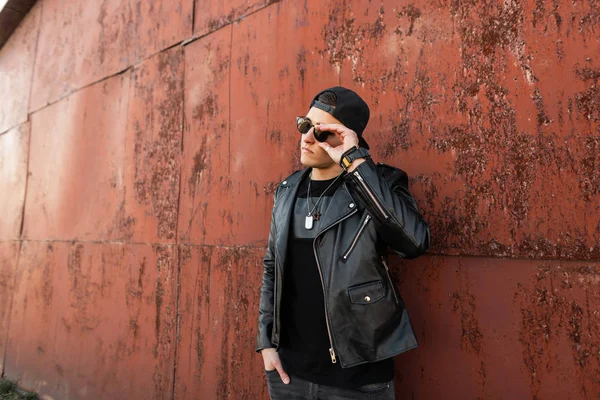 Moderne stijlvolle jonge hipster man in een lederen stijlvolle jas in een modieuze zwarte Baseballpet in trendy donkere zonnebril in Vintage grijze jeans poseren in de buurt van een roestige metalen wand. Amerikaanse kerel buitenshuis. — Stockfoto