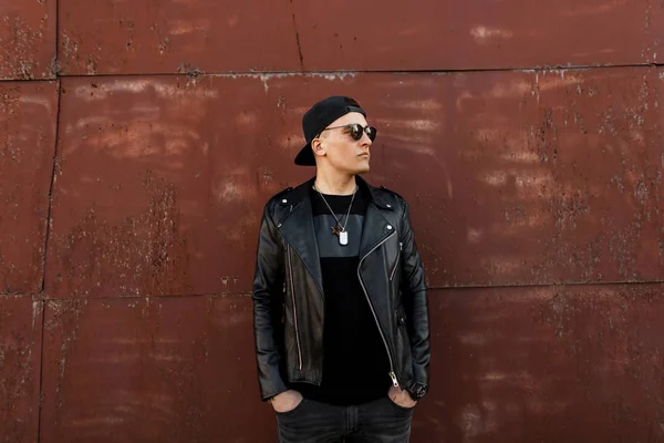 Young American hipster man in a black baseball cap in sunglasses in a leather black jacket in a fashionable t-shirt in gray jeans is standing beside a metal vintage red building. Nice guy outdoors. — Stock Photo, Image