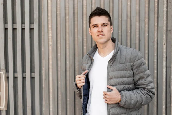 Joven hombre feliz atractivo con un peinado de moda en una chaqueta gris vintage en una elegante camiseta blanca se relaja cerca de una pared de madera vintage de tablones. Europea atractivo chico de vacaciones . — Foto de Stock