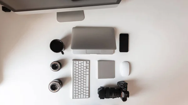 Pohled na bílou plochu, která má notebook, čočky pro fotoaparát, moderní profesionální fotoaparát, černý šálek kávy, klávesnici a telefon. Moderní technologie. — Stock fotografie