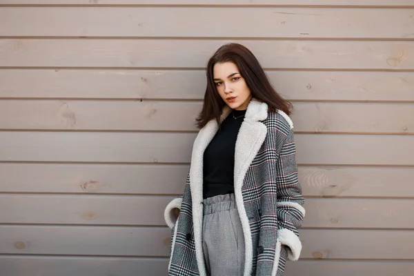 Young pretty beautiful woman in a fashionable black t-shirt in a luxurious plaid jacket in elegant vintage pants is standing on the street near the wooden wall. Attractive girl. Youth style. — Stock Photo, Image