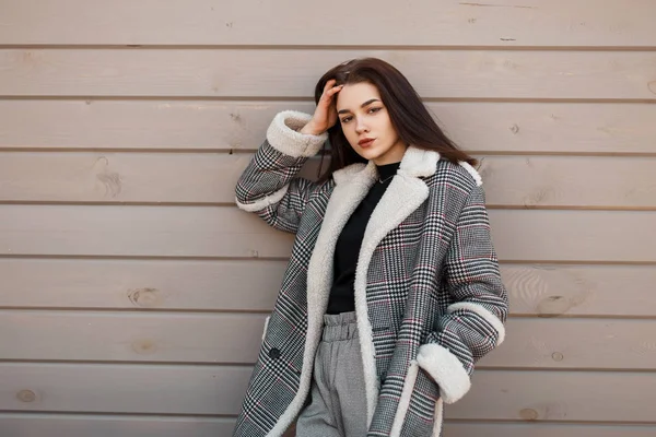 Attraktiv ung brunett kvinna i en trendig t-shirt i en Vintage rutig jacka i grå snygga byxor vilar nära en Vintage trä vägg utomhus. Ganska elegant tjej modell. Vårstil. — Stockfoto