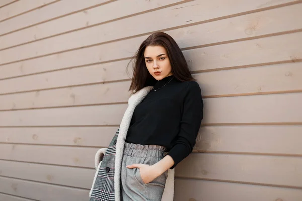 Jolie jeune femme moderne dans un T-shirt noir dans une veste à carreaux à la mode grise dans un pantalon vintage élégant reposant dans la rue près du mur en bois. Belle fille se détend dans le jour du printemps . — Photo