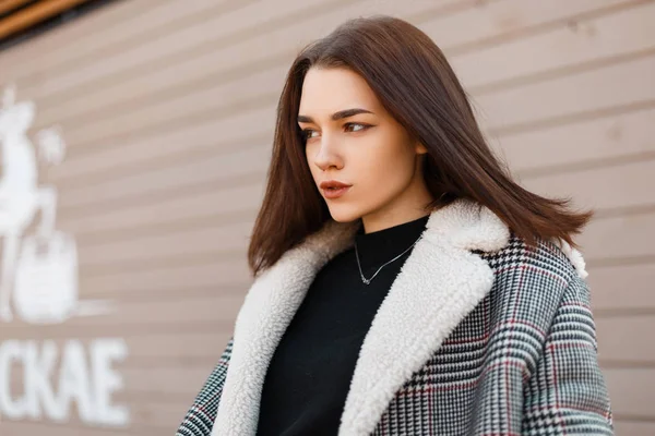 Pretty beautiful young woman model in an elegant spring gray plaid coat in a black trendy T-shirt stands near a modern wooden vintage building on a warm spring day. Attractive cute girl brunette. — Stock Photo, Image