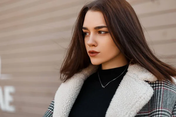 Retrato de uma linda mulher morena bonita com maquiagem natural em uma jaqueta quadriculada na moda com pele em uma camiseta preta elegante perto de uma parede bege de madeira ao ar livre dia de primavera. Menina bonita . — Fotografia de Stock