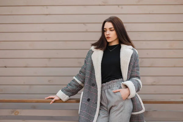 Ganska vacker ung brunett kvinna i en elegant rutig jacka i en svart T-shirt i snygga grå byxor poserar stående nära en beige Vintage träbyggnad. Attraktiv brunett tjej på promenaden — Stockfoto