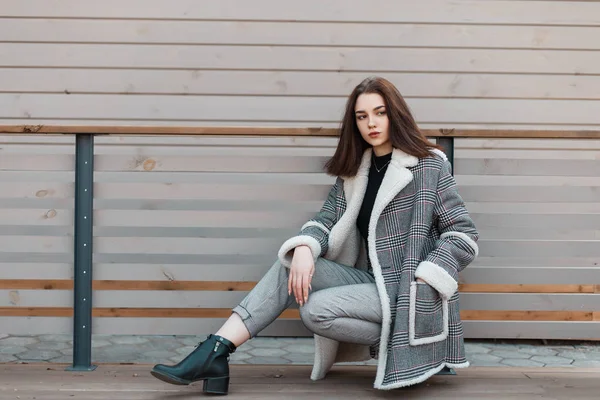 Mulher bonita e elegante em calças cinza na moda em sapatos de couro preto em uma jaqueta quadriculada elegante posando sentado perto de um edifício moderno de madeira na cidade. bonito atraente menina relaxa . — Fotografia de Stock