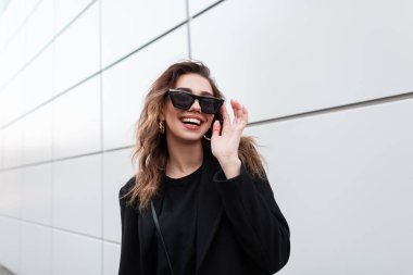 Gri bir binanın yakınında açık havada poz moda siyah ceket bir gülümseme ile şık güneş gözlüğü Pretty neşeli genç hipster kadın. Kentsel Avrupa pozitif kız gülüyor.