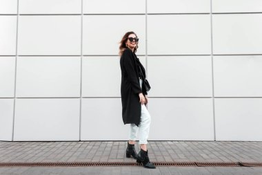 Bir çanta ile deri ayakkabı vintage kot şık bir uzun ceket trendy güneş gözlüğü Olumlu güzel genç hipster kadın açık havada beyaz modern duvar yakınında duruyor. Kentsel mutlu kız modeli.