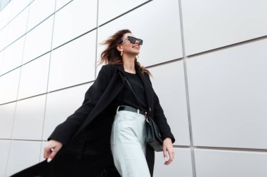 Siyah trendy giysiler içinde güneş gözlüğü olumlu bir gülümseme ile Modern neşeli genç hipster kadın bir bahar gününde şehir etrafında seyahat. Neşeli şehirli kız hafta sonu zevk. Sokak modası.