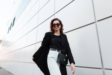 Positive young stylish hipster woman in sunglasses in an elegant coat in a T-shirt in vintage jeans with a leather handbag is walking and smiling near a modern building in the city.Cheerful girl walks clipart