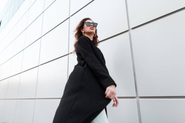 Eski moda güneş gözlüklü, eski moda siyah ceketli genç ve güzel hippi kadın, sıcak bir bahar gününde modern bir binanın yakınına gider. Şık, çekici bir kız şehirde geziyor. Retro biçimi.