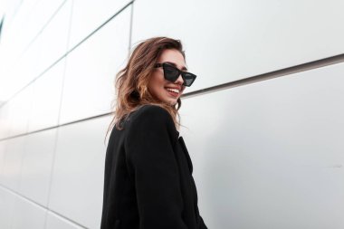 Siyah zarif ceket güzel bir gülümseme ile koyu güneş gözlüğü Pretty neşeli genç hipster kadın sıcak bir bahar gününde gri duvarın yanında duruyor. Pozitif mutlu kız modeli hafta sonu sahiptir. 